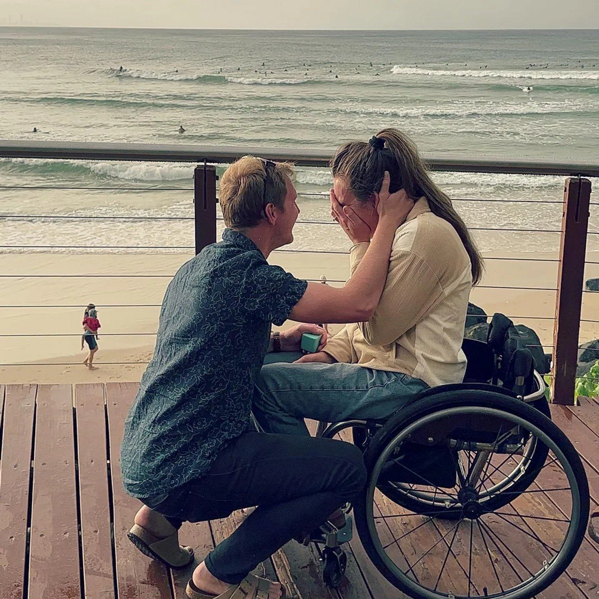 A man wearing a short sleeve shirt, jeans and sandals proposing to a woman wearing jeand and in a wheelchair on a boardwalk above a beach.