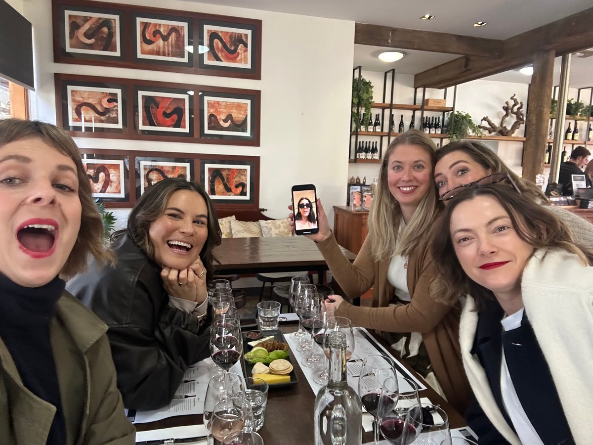 A selfie of five women smiling at a dinner table while one holds a phone with a woman on video call.
