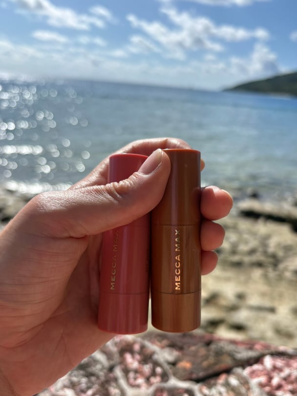 Zoe holds the Mecca Max contour and blush sticks in front of the ocean.