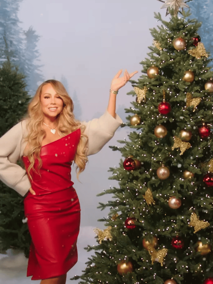 Mariah Carey wearing a long red gown posing next to her Christmas tree. 