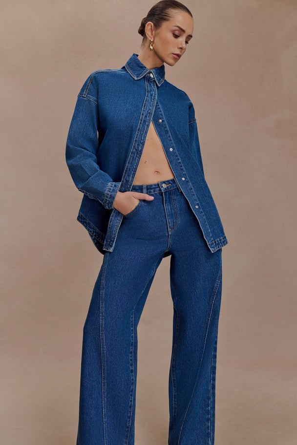 Woman wearing an oversized, long-sleeved denim shirt with matching wide-leg paneled denim jeans from Meshki.