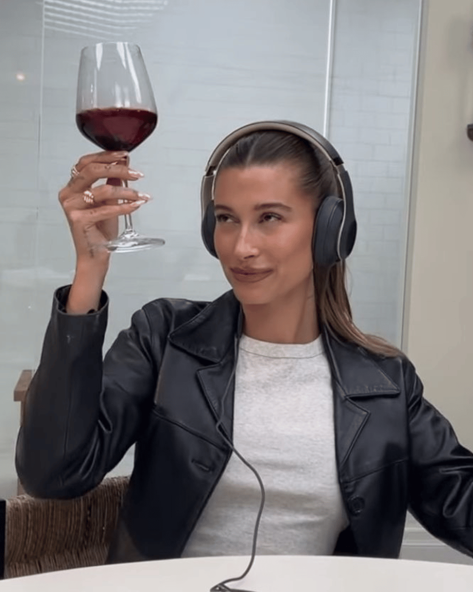 Hailey Bieber wearing a pair of headphones and cheersing a glass of red wine.