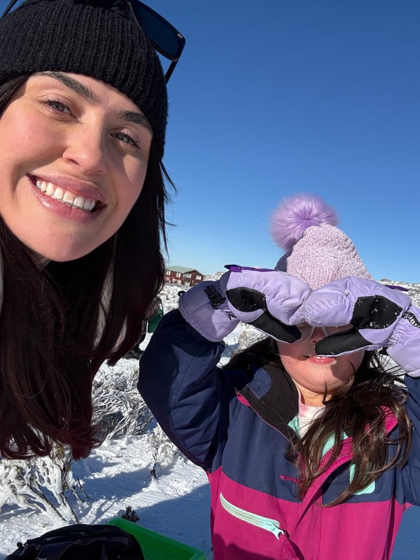Selfie of Nikki smiling at the snow with her daughter making heart hands.