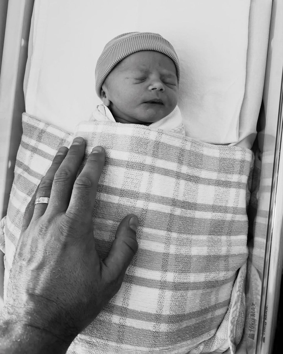 Olympia Valance and Thomas Bellchambers have welcomed a baby boy, Billy Bellchambers, who is swaddled wearing a beanie as Bellchambers' hand rests on top of the blanket.