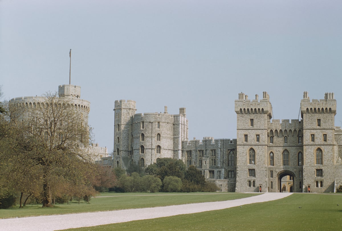 Windsor Castle on a sunny day in England. It has turrets and a grand entrance lined by grass and trees.