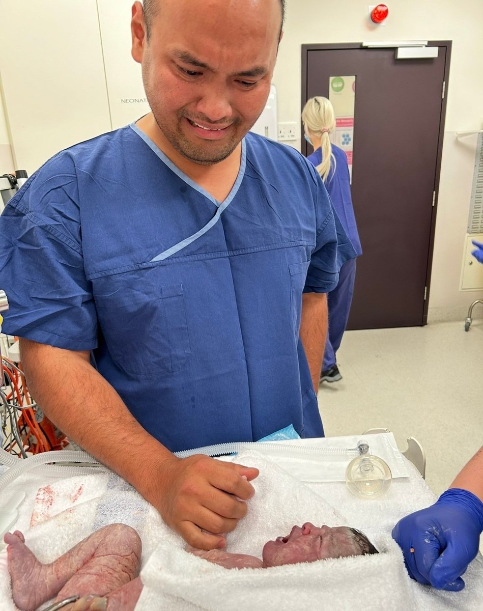 Sabrina's husband wears blue hospital scrubs as he cries holding his newborn daughter's hand.