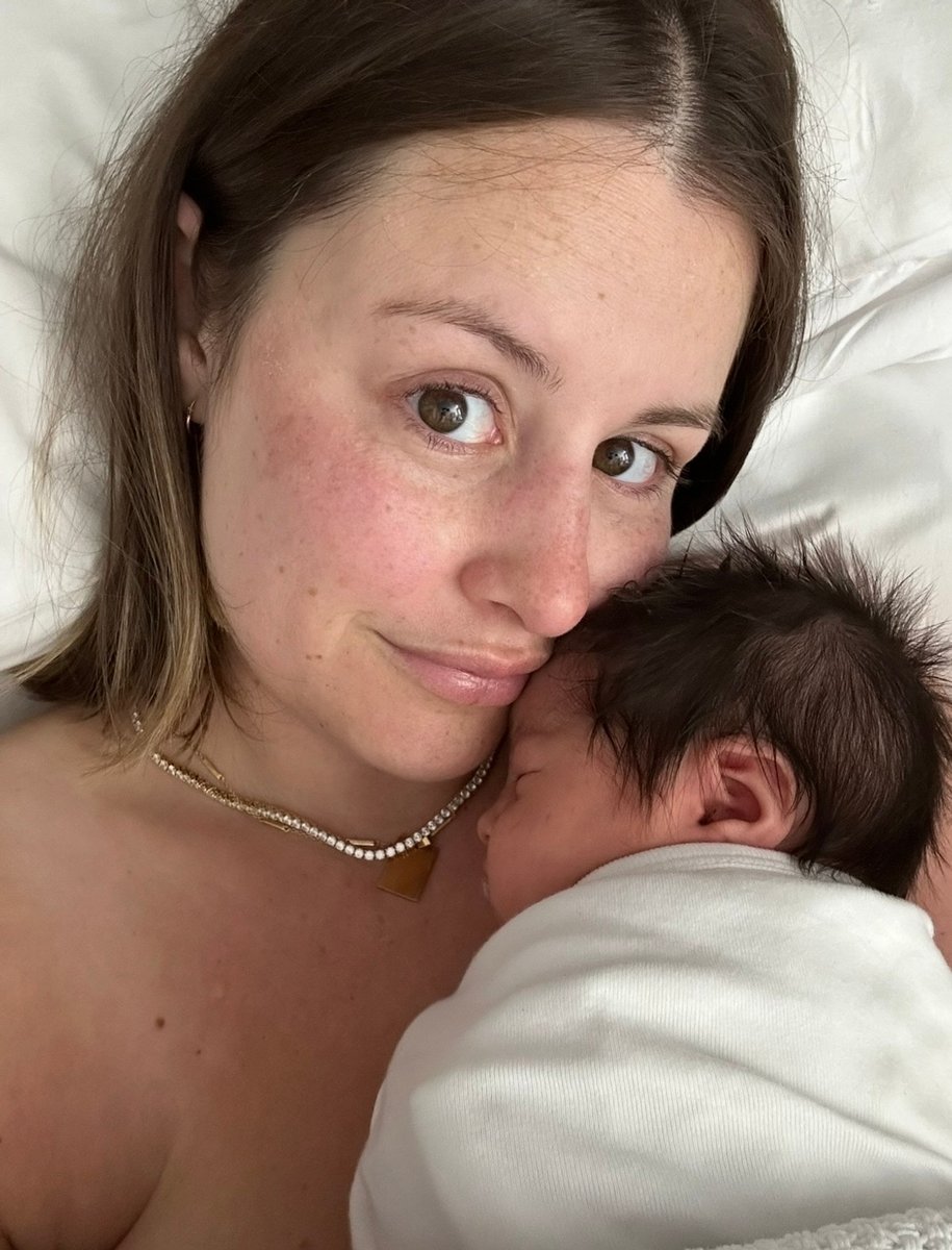 Sabrina smiles with her baby Sienna swaddled in a white blanket on her chest.