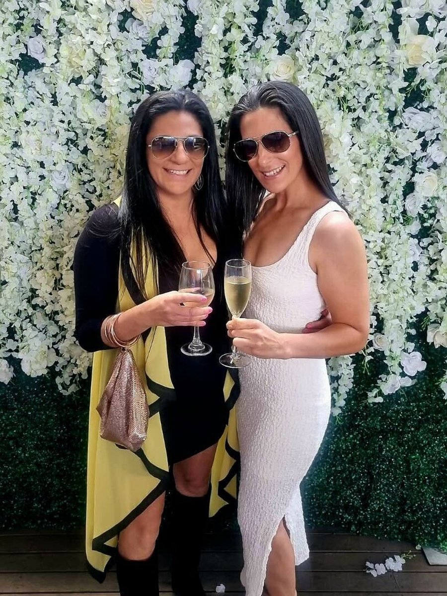 Sarah Hussein wears a black and yellow dress and holding a glass of wine next to a woman in a white dress holding a glass of champagne as they stand in front of a white wall of flowers.