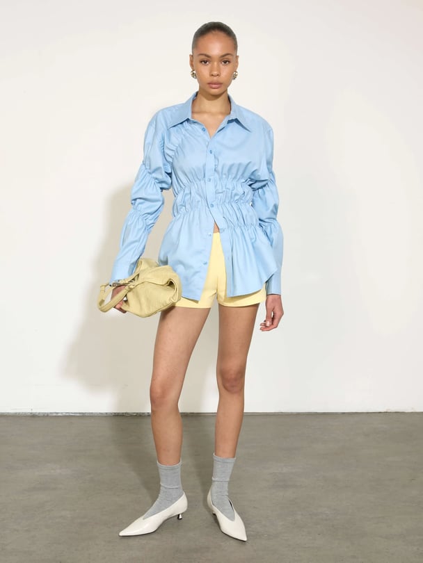 Model in blue button up with scrunch detailing, yellow short and white kitten heels. 