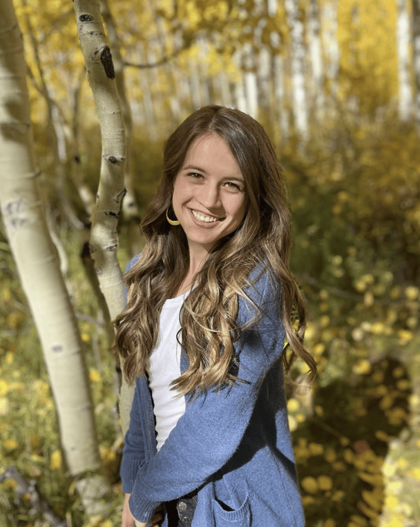 Ruby Franke's daughter, Shari Franke smiles wearing a white t-shirt and blue cardigan in front of trees.