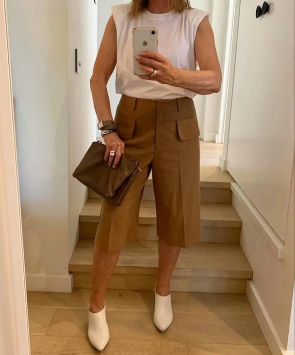 Middle aged woman wearing white shoulder pad tee, camel tailored Bermuda shorts and white heeled boots.