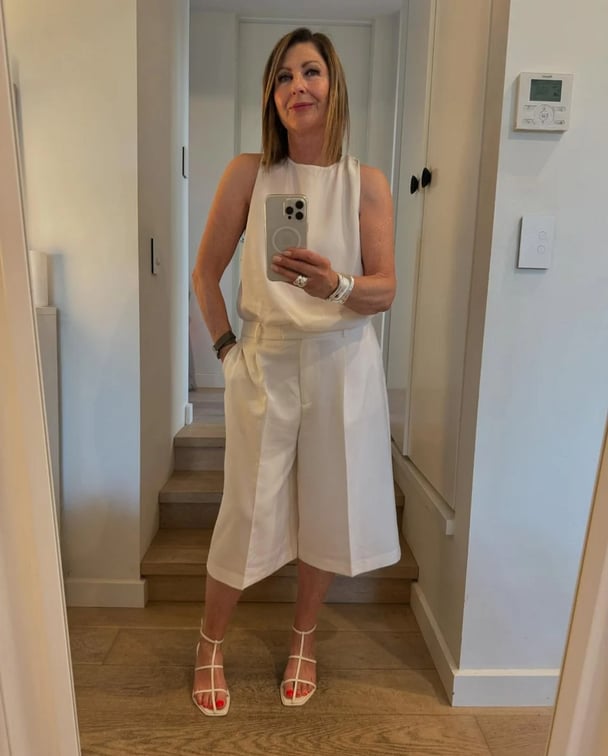 Middle aged woman wearing a white singlet, white Bermuda shorts and white strappy sandals.