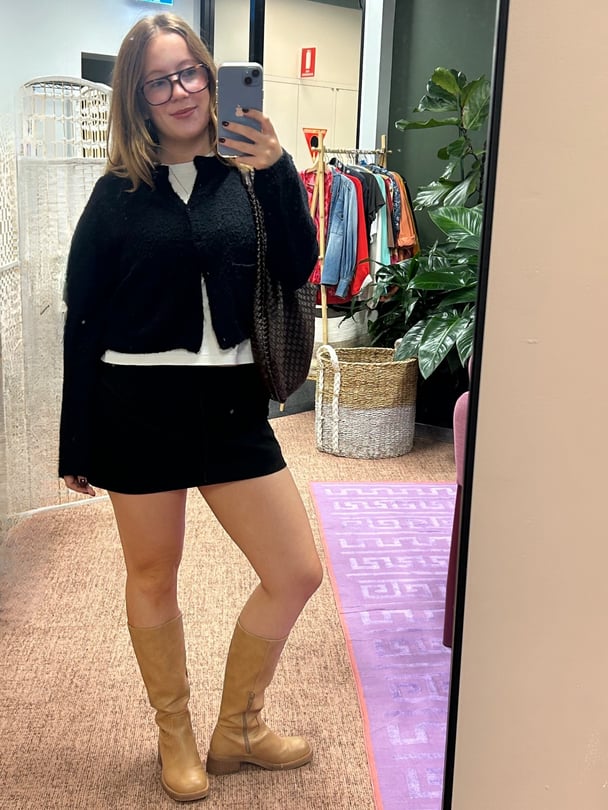 Mirror selfie of Lucy in boots with a black miniskirt and black cardigan.