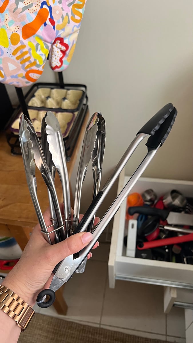 Hand holding four pairs of tongs, with a messy kitchen drawer in the background.