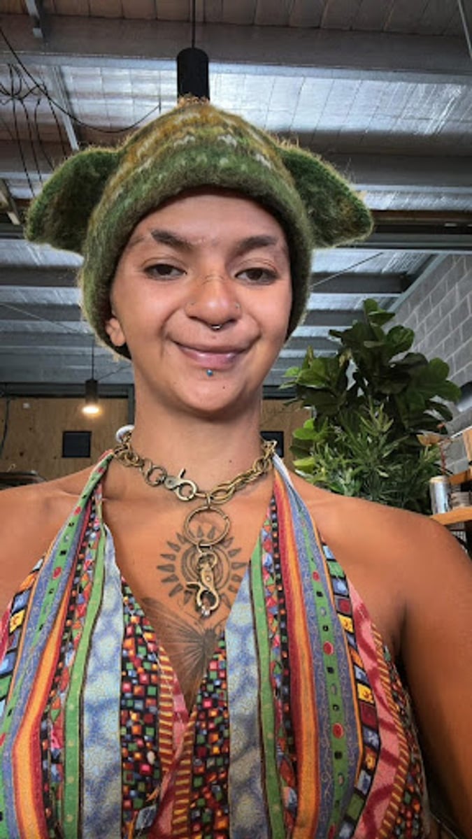 Photo of Josephine Hewitt. She is wearing a knitted beanie with ears, has facial tattoos and multiple piercings, a visible chest tattoo, and is wearing an industrial chain necklace.